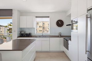 A modern kitchen with white cabinetry, a built-in oven, a coffee machine, and a window with a view of buildings. - Lovely 3BR Penthouse in Shenkin area by HolyGuest (Tel Aviv-Yafo)