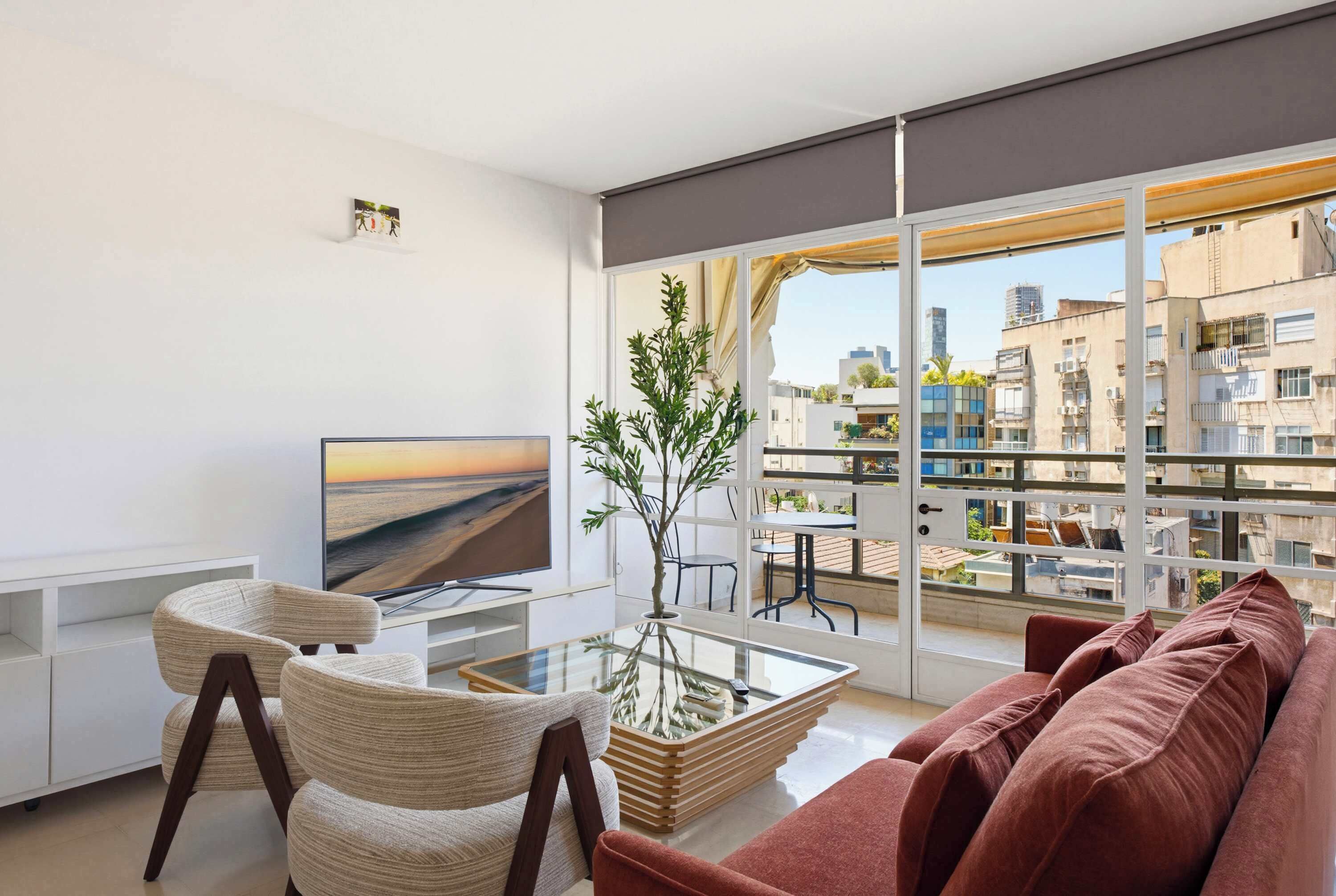 A modern living room with a sofa, chairs, a TV, a glass coffee table, and a balcony with a city view.