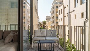 Un balcon doté d’un canapé et d’une table, entouré d’une rambarde en métal, d’où l’on peut admirer une rangée d’immeubles.