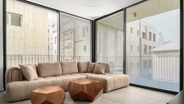 Un salon moderne avec un canapé d’angle, deux tables basses hexagonales en bois et de grandes fenêtres du sol au plafond offrant une vue sur la ville.