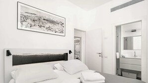 Une chambre d’hôtel moderne, avec un lit, une photographie encadrée accrochée au mur, une salle de bain dotée d’un miroir et d’un lavabo, et une porte donnant sur une autre pièce.