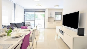 A modern kitchen with a large TV, a dining area with a table set for two, and a view through the glass door. - Garden Kalisher 3BR near the Beach by HolyGuest (Tel Aviv-Yafo)