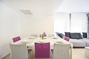Un coin repas avec une table blanche dressée pour quatre personnes, des chaises violettes et un canapé en arrière-plan.