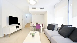 A modern living room with a flat-screen TV, a sofa, a coffee table, and a dining area with chairs and a table. - Garden Kalisher 3BR near the Beach by HolyGuest (Tel Aviv-Yafo)