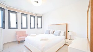 A modern bedroom with a large bed, a bedside table, a chair, and a window. - Garden Kalisher 3BR near the Beach by HolyGuest (Tel Aviv-Yafo)