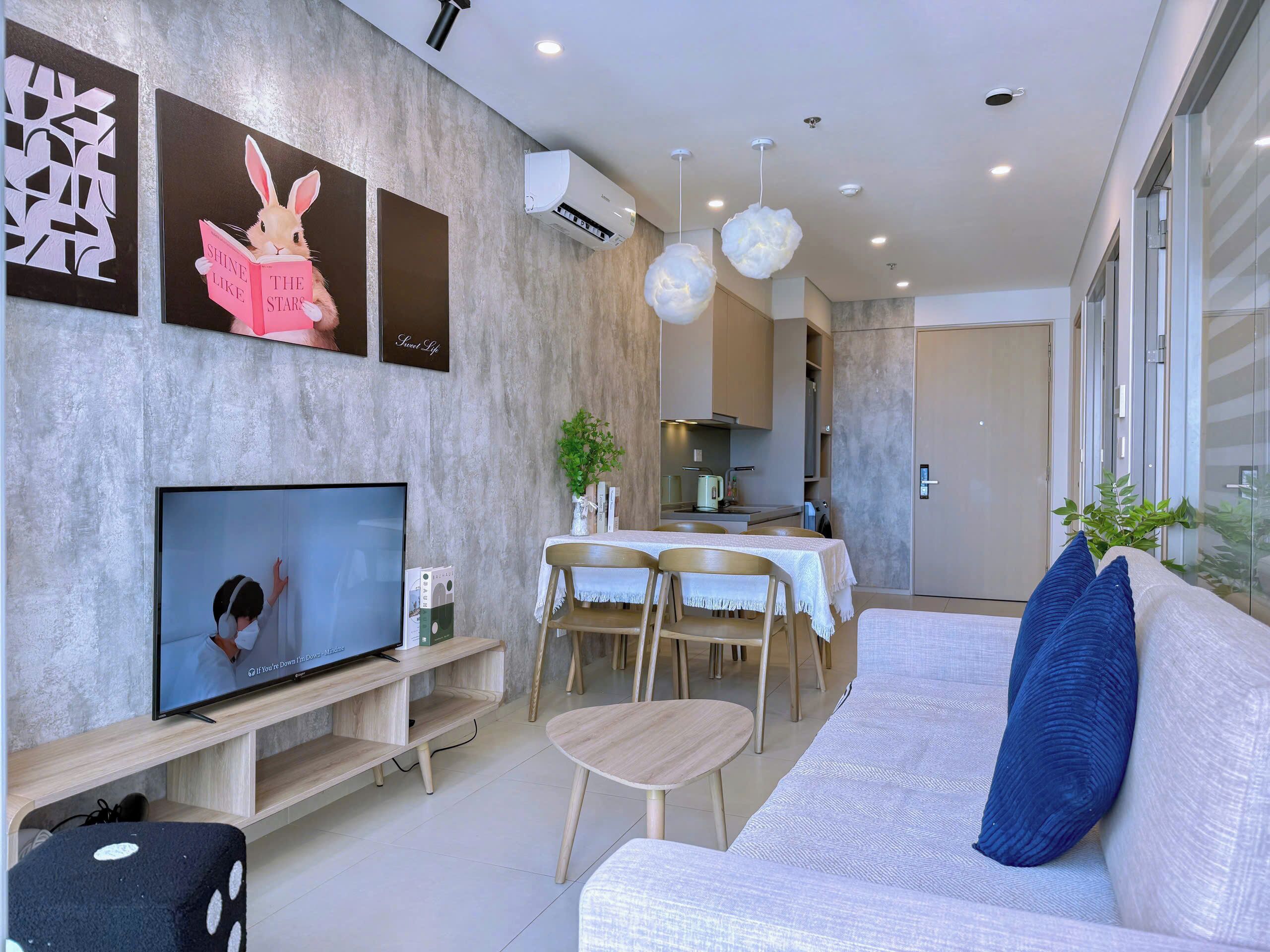 A modern living room with a flat-screen TV, a sofa with a blue pillow, a dining area with a table and chairs, and a kitchen in the background.