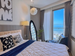 A hotel room with a large bed, a view of the city, a potted plant, and a modern lamp.