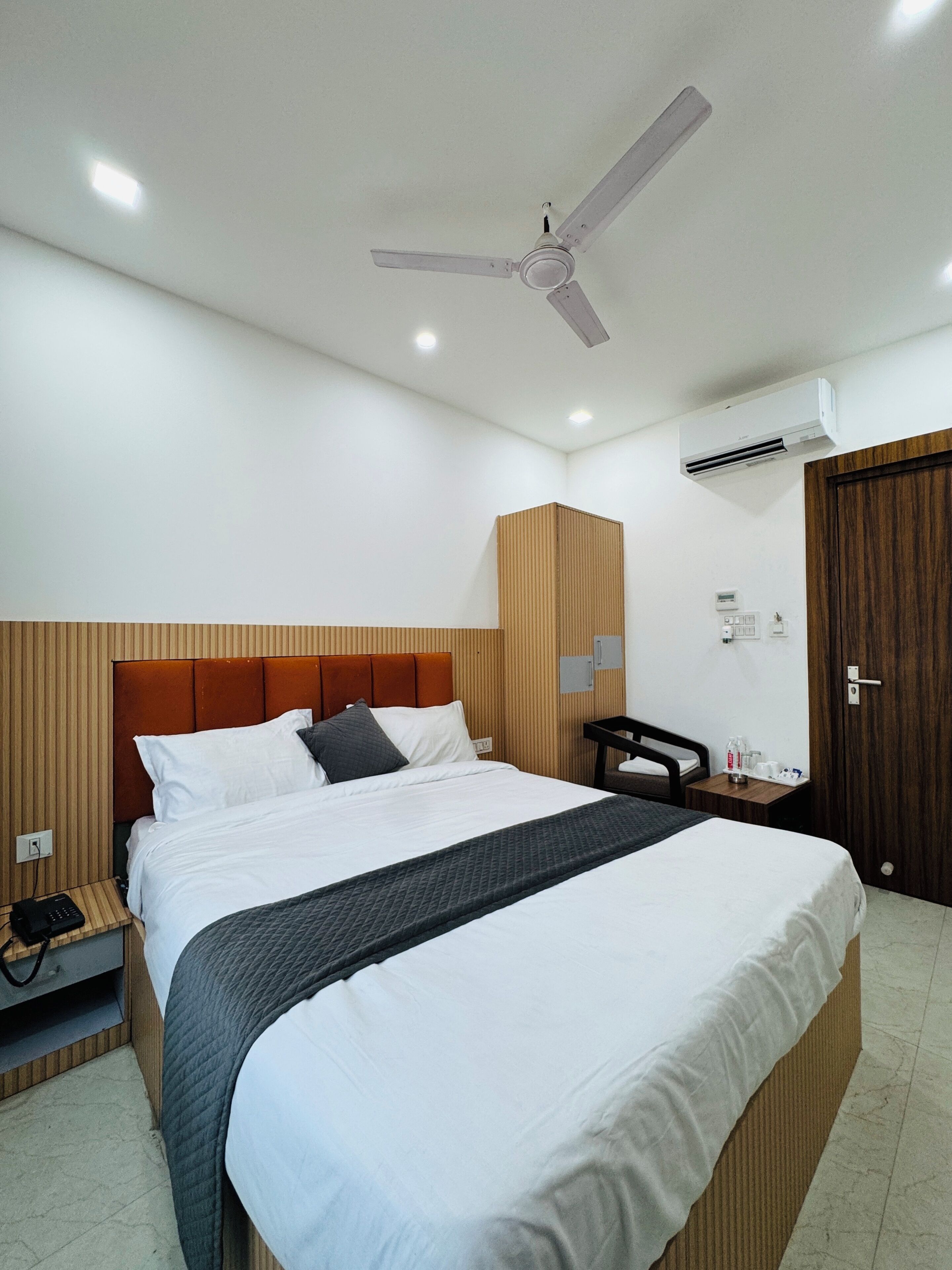 A hotel room with a bed, a nightstand, a desk, a chair, a wardrobe, a ceiling fan, and an air conditioning unit.