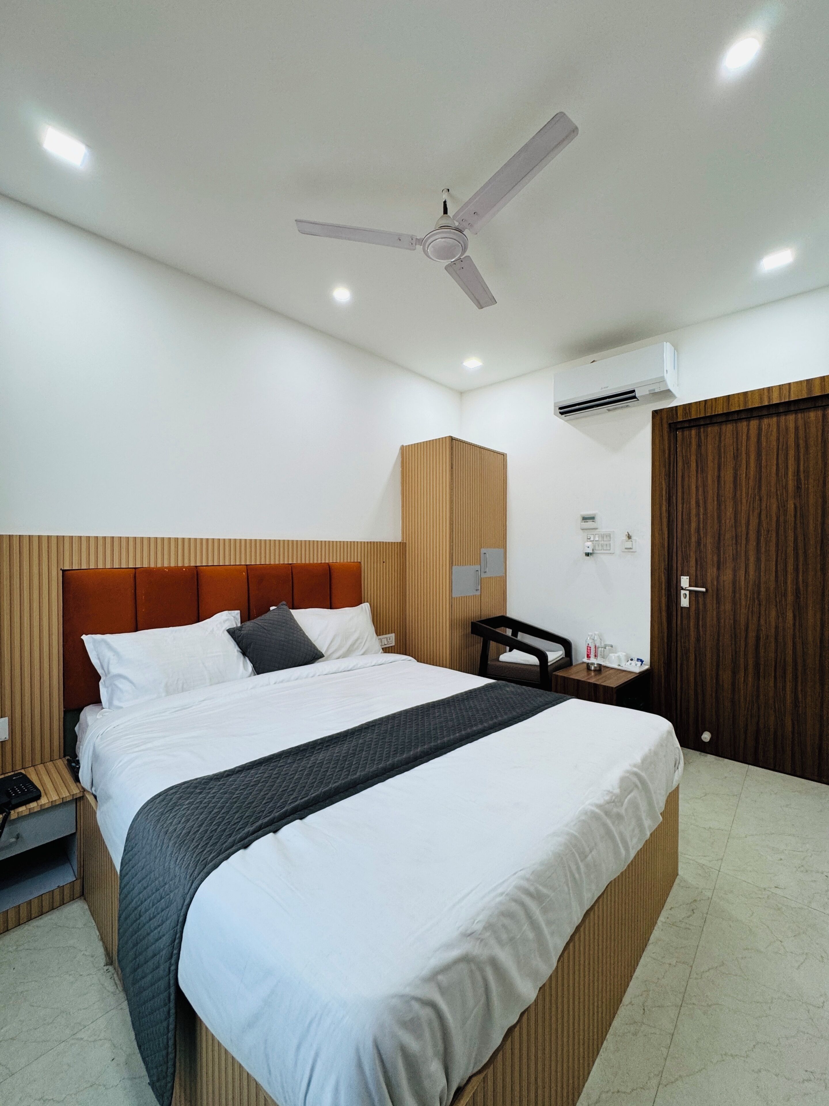 Una habitación de hotel con cama, ventilador de techo, aire acondicionado, armario de madera, mesita de noche con lámpara y un atril con teléfono.