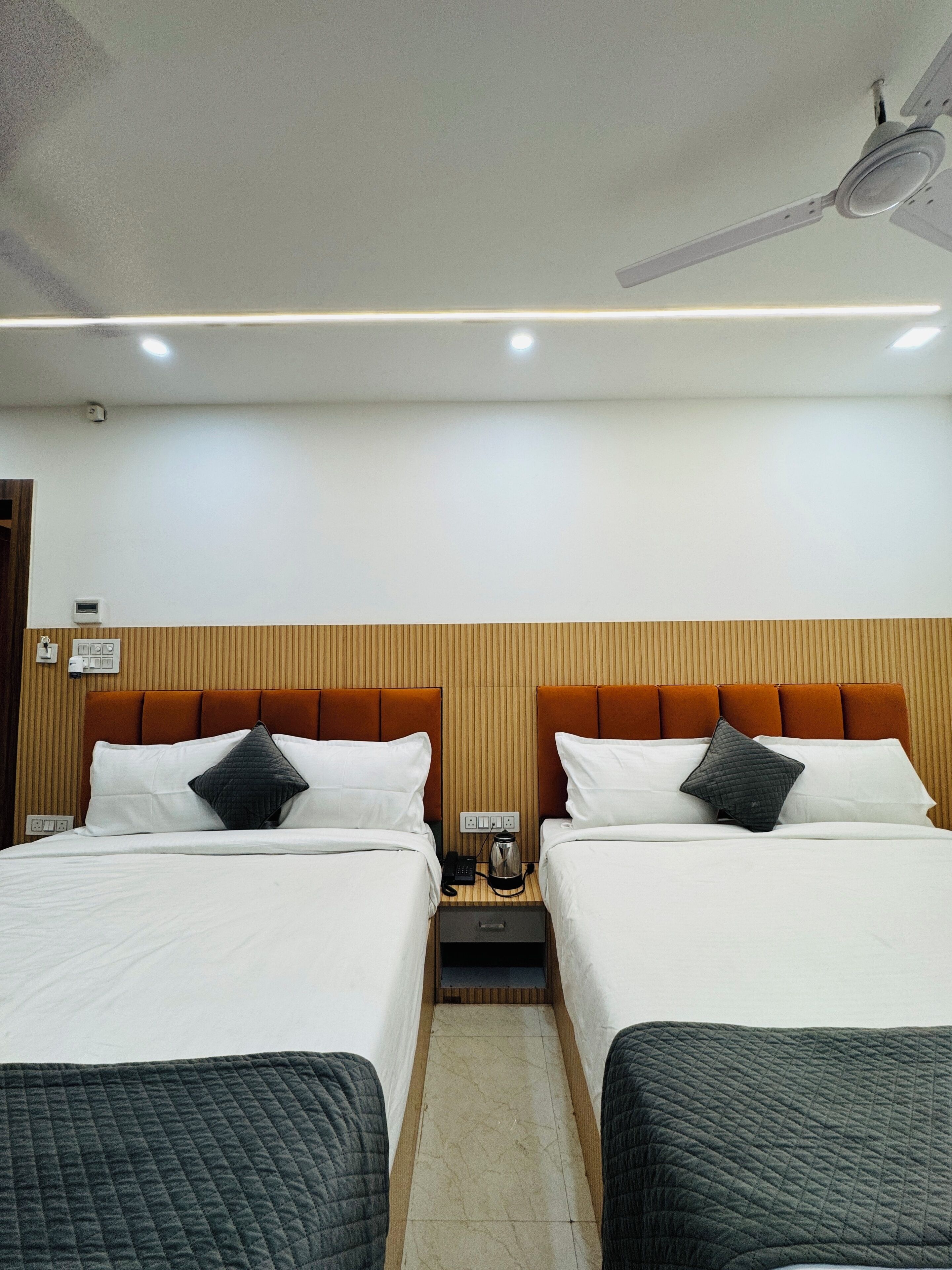 A hotel room with two beds, a wooden headboard, a ceiling fan, and a bedside table with a phone and a kettle.