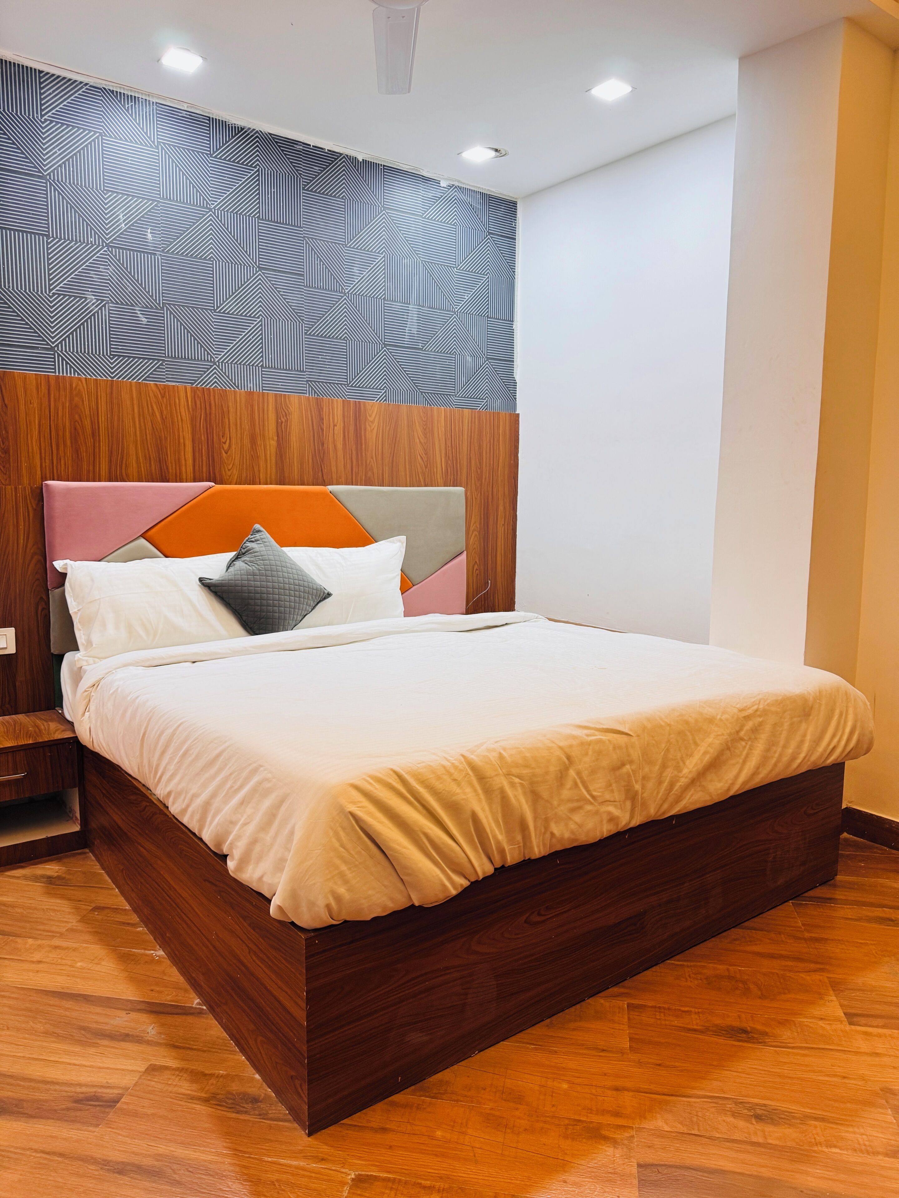 Une chambre moderne avec une tête de lit en bois, un lit recouvert d’une couvre-lit beige et un mur orné d’un motif géométrique.