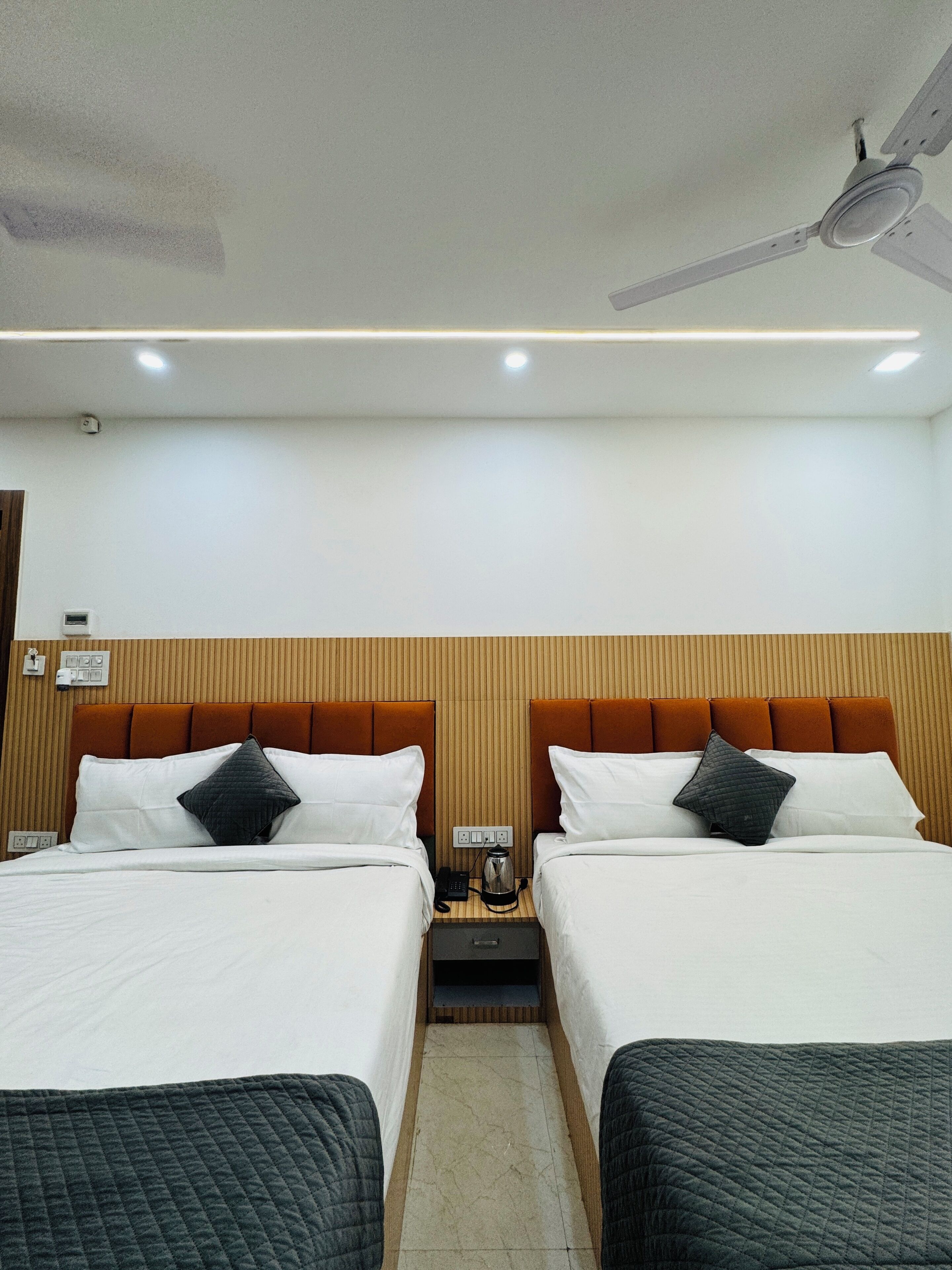 A hotel room with two beds, a wooden headboard, a nightstand with a phone and a kettle, and a ceiling fan.