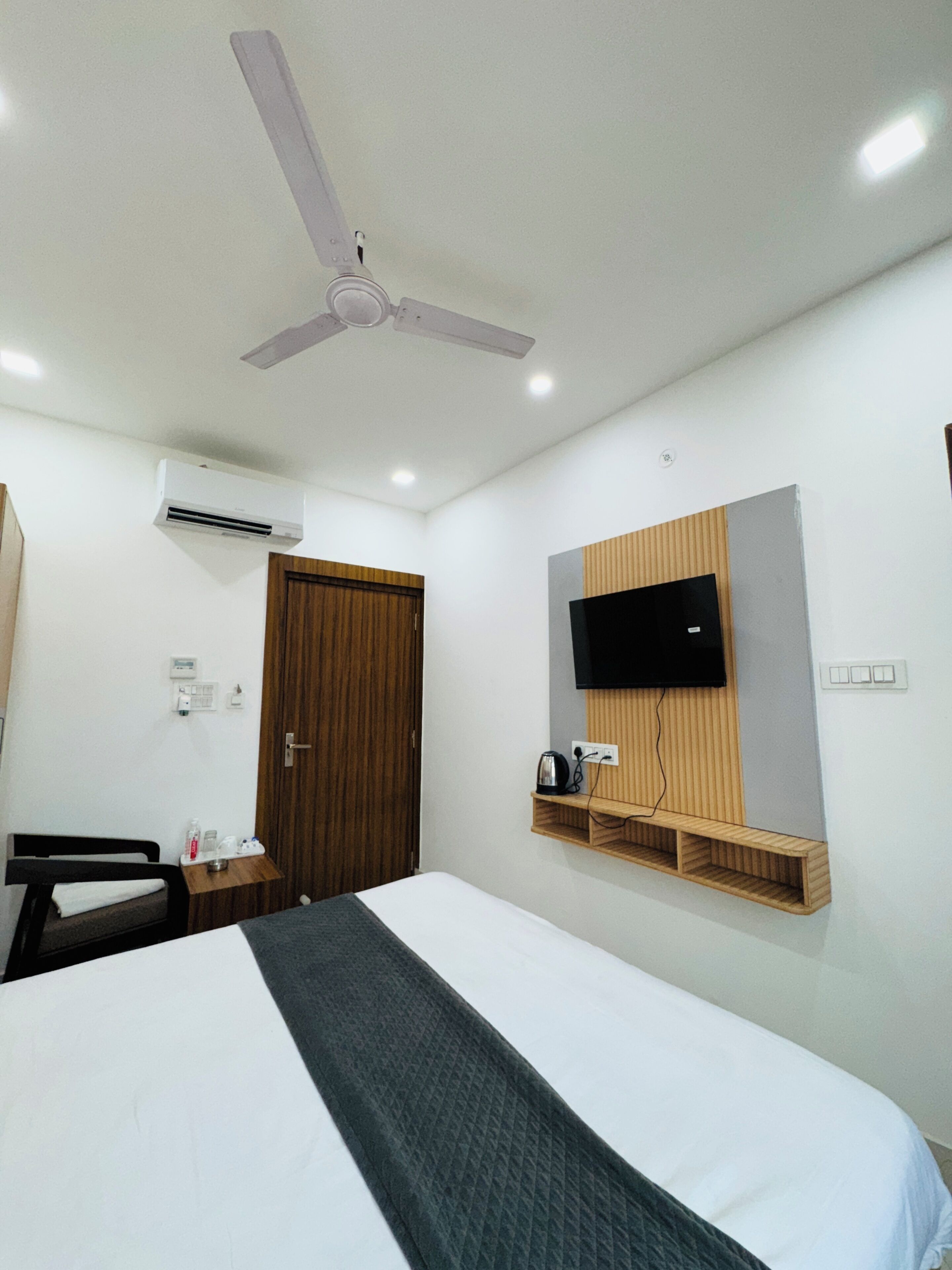 A hotel room with a bed, a TV mounted on the wall, a ceiling fan, an air conditioning unit, a wooden door, and a small wooden shelf with a kettle.