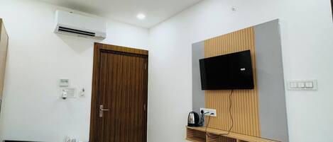 A hotel room with a bed, a TV mounted on the wall, a ceiling fan, an air conditioning unit, a wooden door, and a small wooden shelf with a kettle.