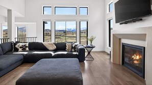 Een moderne woonkamer met een hoekbank, een flatscreen televisie, een open haard en grote ramen met uitzicht op de bergen.