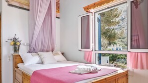 Ein Schlafzimmer mit einem hölzernen Bett, einer rosa Bettdecke, einem Fenster mit Vorhängen und Blick ins Grüne.