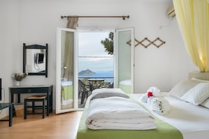 A hotel room with a bed, a desk, a chair, a small table, and a view of the sea through an open door. - Room 14 at Pagali Hotel with Sea View (Órmos Aiyialís)