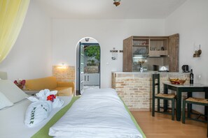 Een kamer met een bed, een groen gestreept vloerkleed, een houten eettafel met twee stoelen, een keuken met een bakstenen muur en een uitzicht door een boog.