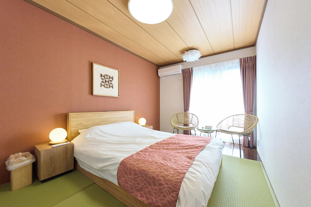 A hotel room with a bed, bedside lamps, a wall-mounted air conditioner, a window with curtains, and a chair by the window.