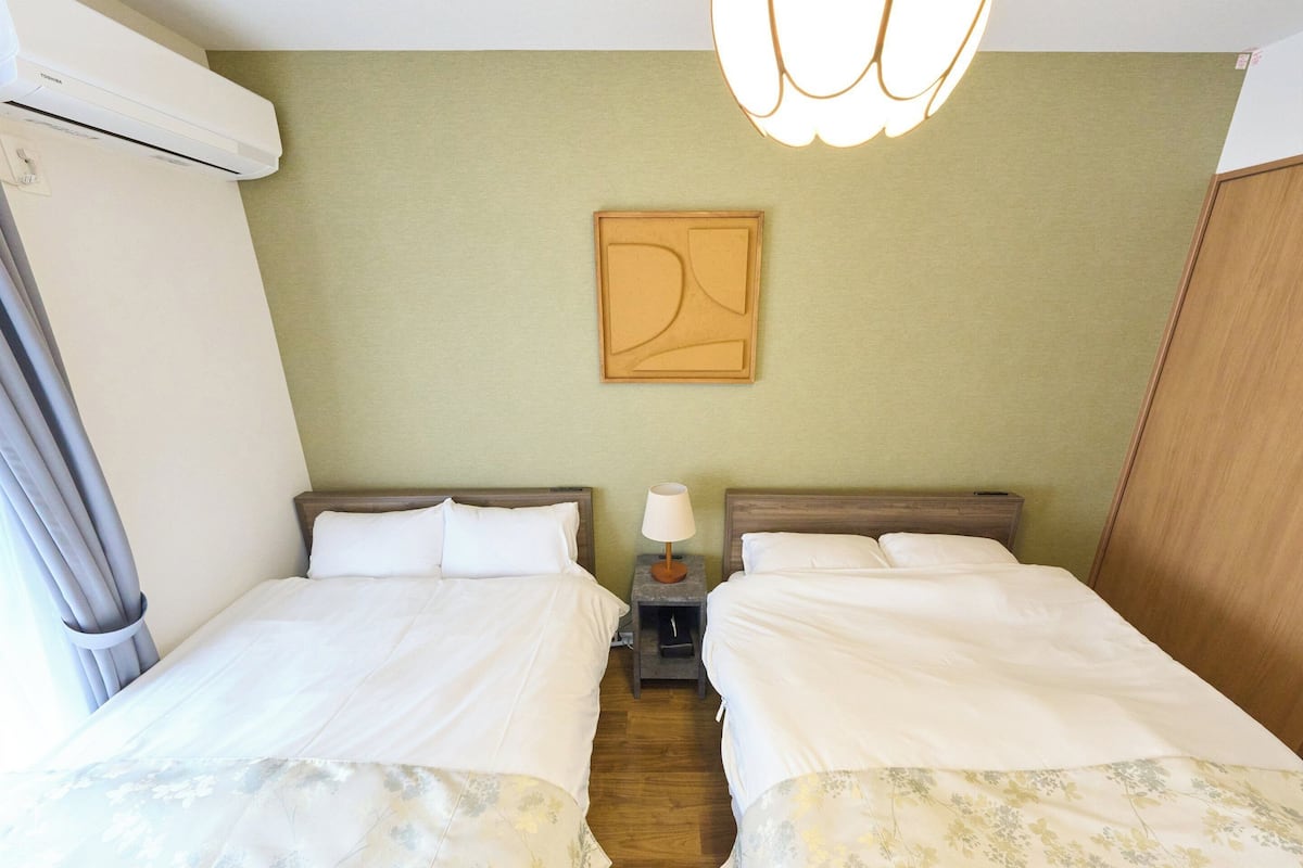 A hotel room with two beds, a nightstand with a lamp, a wall-mounted air conditioner, and a painting.