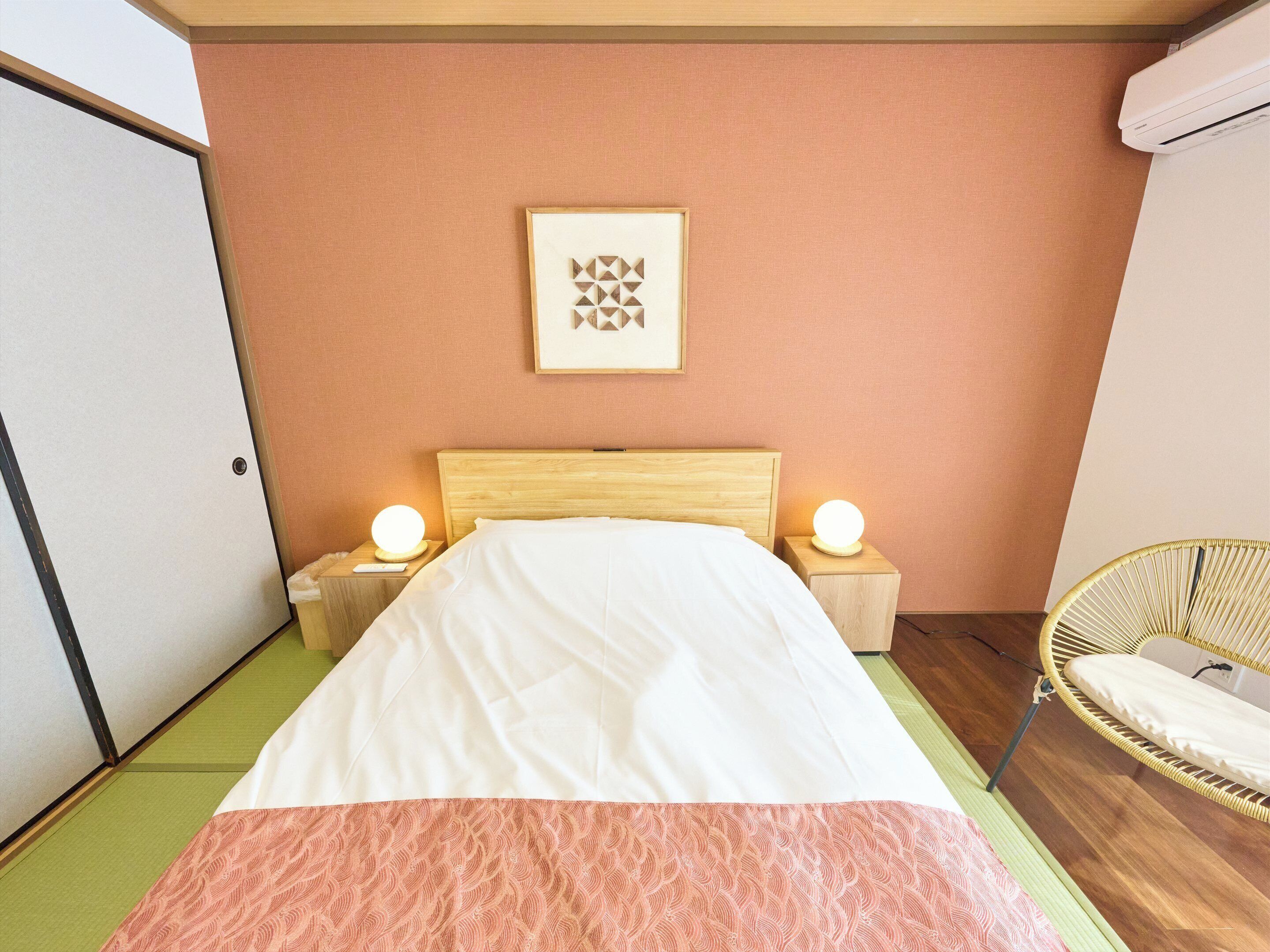 A neatly made bed with a patterned blanket, two bedside lamps, a framed picture on the wall, and a wicker chair.
