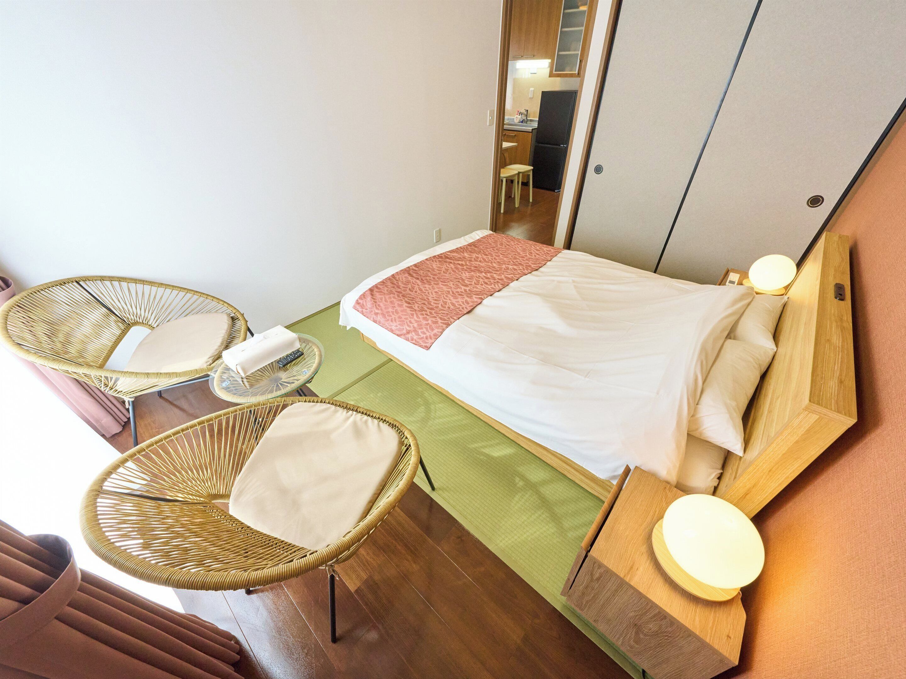 A hotel room with a bed, two wicker chairs, a small table, and a wooden nightstand with a lamp.