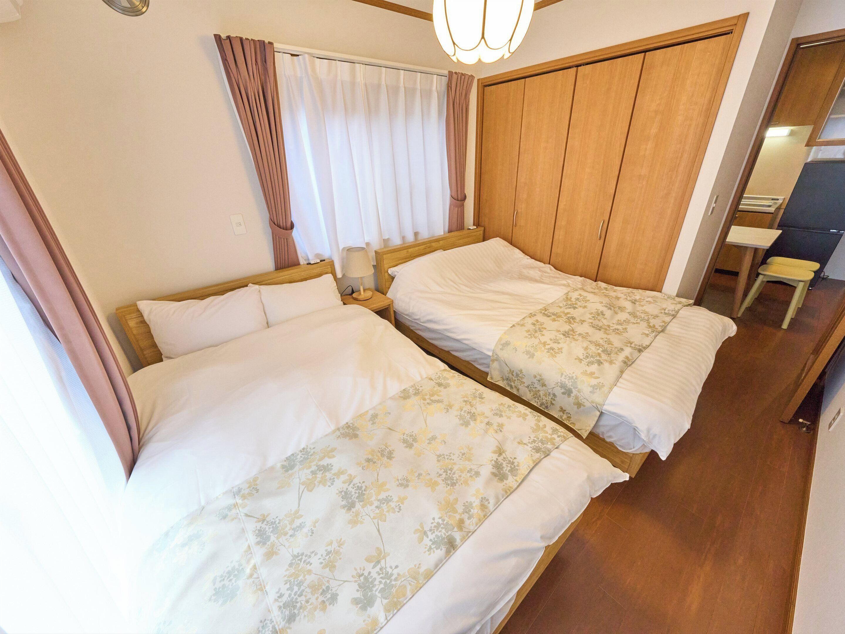 A hotel room with two beds, wooden headboards, a wooden wardrobe, a small desk, and a chair.