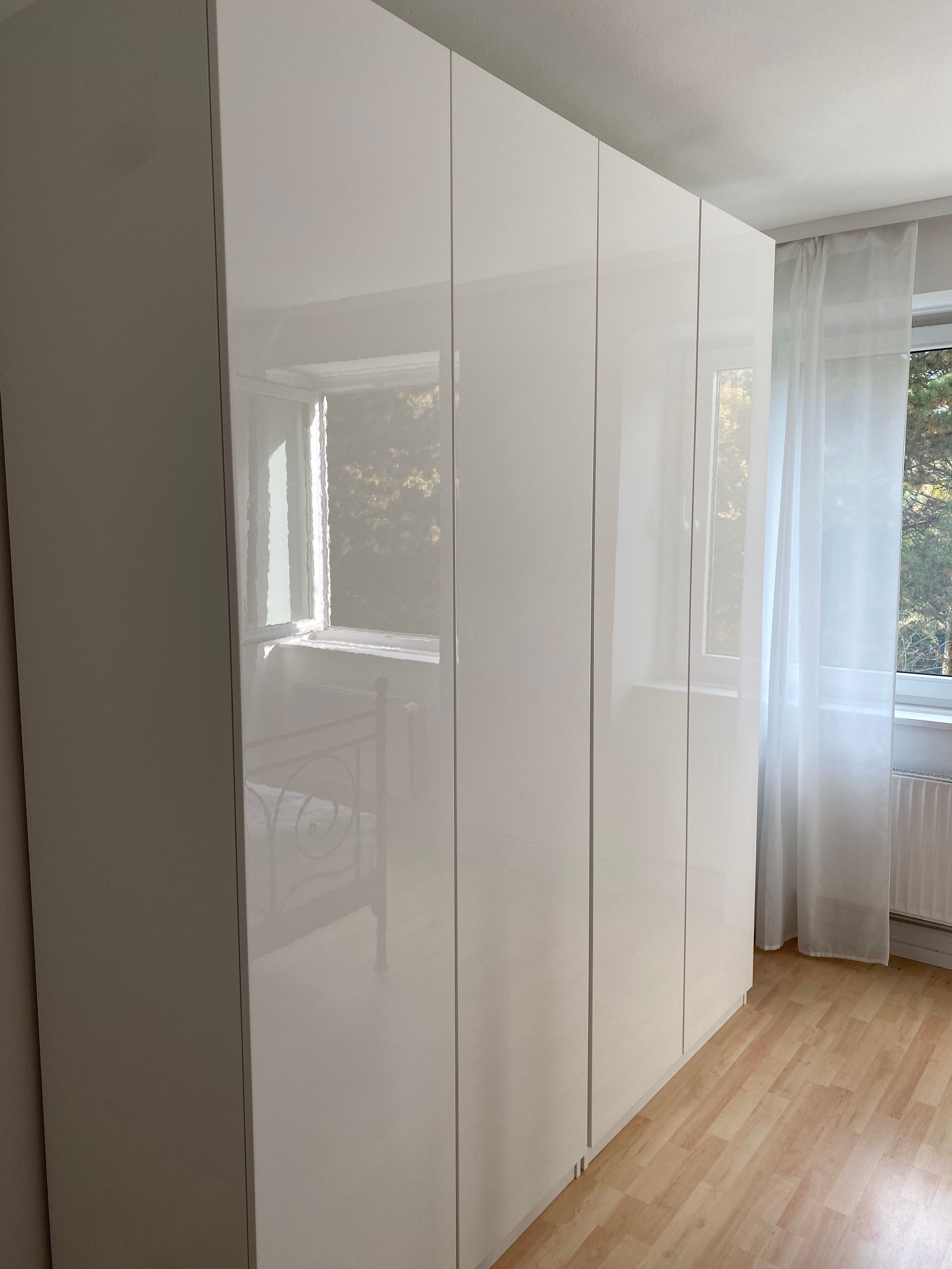 Ein Zimmer mit einem großen, weißen Kleiderschrank, Holzboden und einem Fenster mit transparenten Vorhängen.