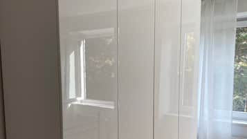 Ein Zimmer mit einem großen, weißen Kleiderschrank, Holzboden und einem Fenster mit transparenten Vorhängen.