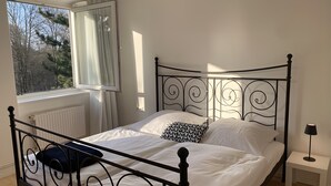 A bed with a metal frame, white bedding, a patterned pillow, a nightstand with a lamp, and a window with curtains. - Cozy Apartment in Hannover – Near City, Fair, MHH with Balcony and Wi-Fi (Hannover)