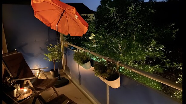 A balcony with a chair, an orange umbrella, hanging planters, and a view of trees. - Cozy Apartment in Hannover – Near City, Fair, MHH with Balcony and Wi-Fi (Hannover)