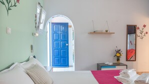 Een hotelkamer met een bed, een bureau, een spiegel, een blauwe deur en een plank met bloemen.