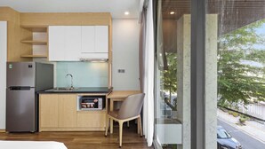 Uma cozinha compacta com geladeira, pia e armários de madeira, uma pequena área de refeições com uma cadeira, e uma grande janela com vista para a rua.