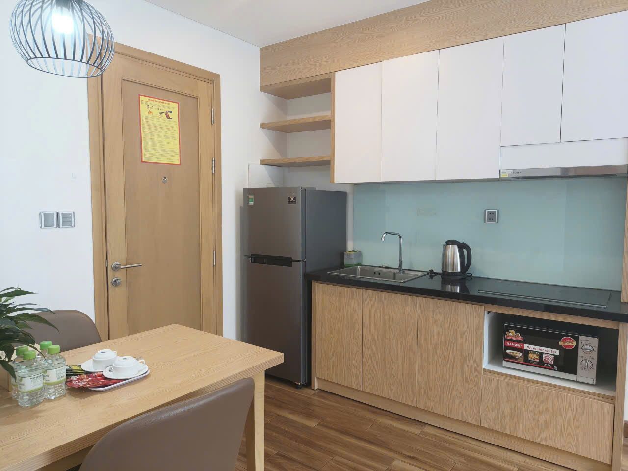 Una cocina compacta con refrigerador, pileta y microondas. Hay un armario de madera sobre la pileta y una pequeña estantería con una caja.