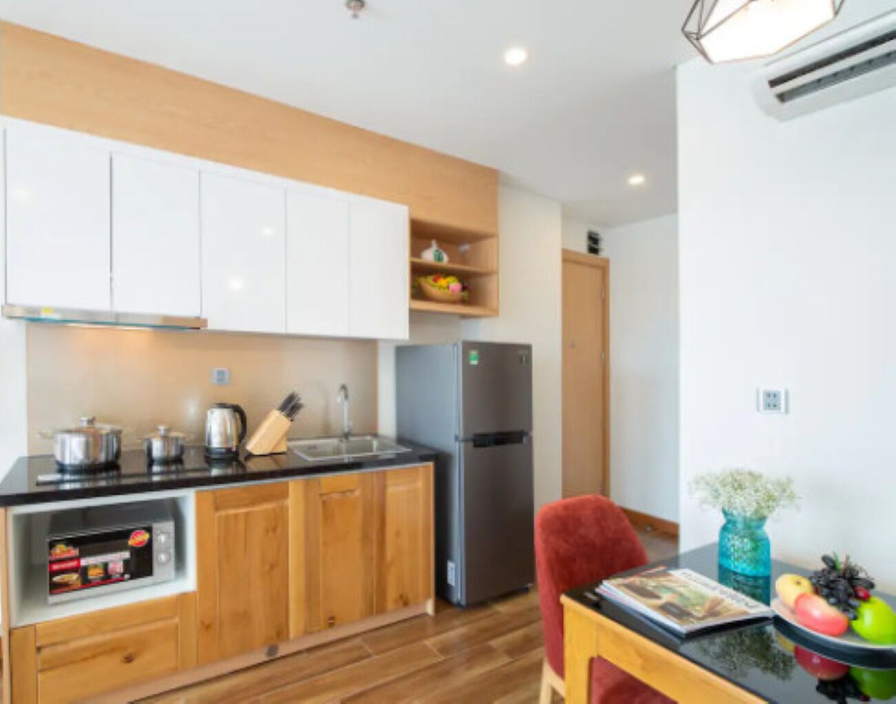 A compact kitchen with wooden cabinets, a stainless steel refrigerator, a built-in oven, and a dining area with a table and chair.