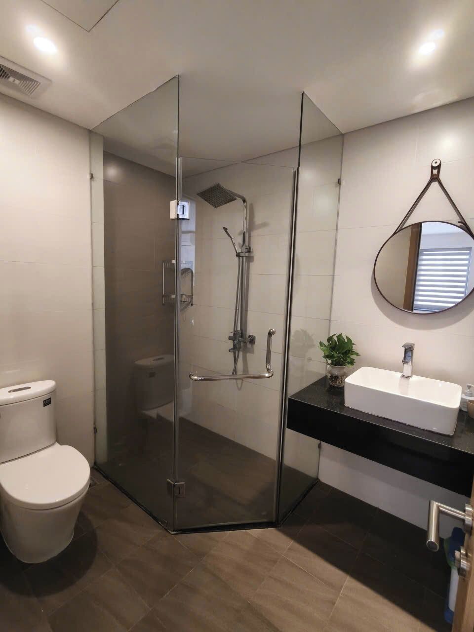 A modern bathroom with a glass-enclosed shower, a white toilet, a black countertop, and a round mirror.