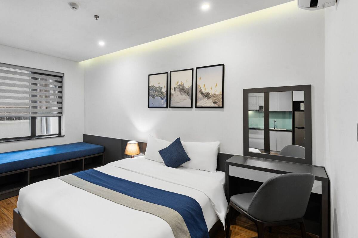A modern hotel room with a bed, bedside table, chair, and a view of the kitchen.