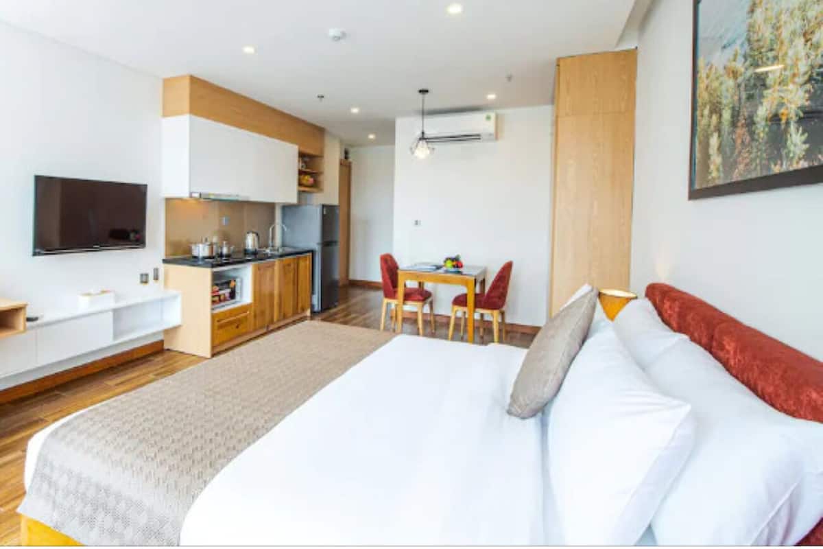 A modern hotel room with a large bed, a dining table, a kitchen area, and a TV.