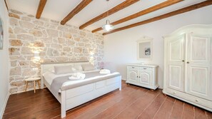 A bedroom with a stone wall, wooden beams, a bed with white bedding, a nightstand, a dresser, and a wardrobe.