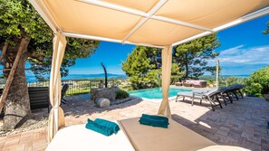 A covered outdoor area with a pool, lounge chairs, and a view of the sea.