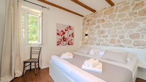 A bedroom with a stone wall, a bed with white linens, a wooden chair, and a window with curtains. - Vila Pantovcak with stunning views by Villas Guide (Mundanije)