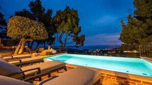 A swimming pool with lounge chairs, a stone wall, and a view of the sea at dusk.