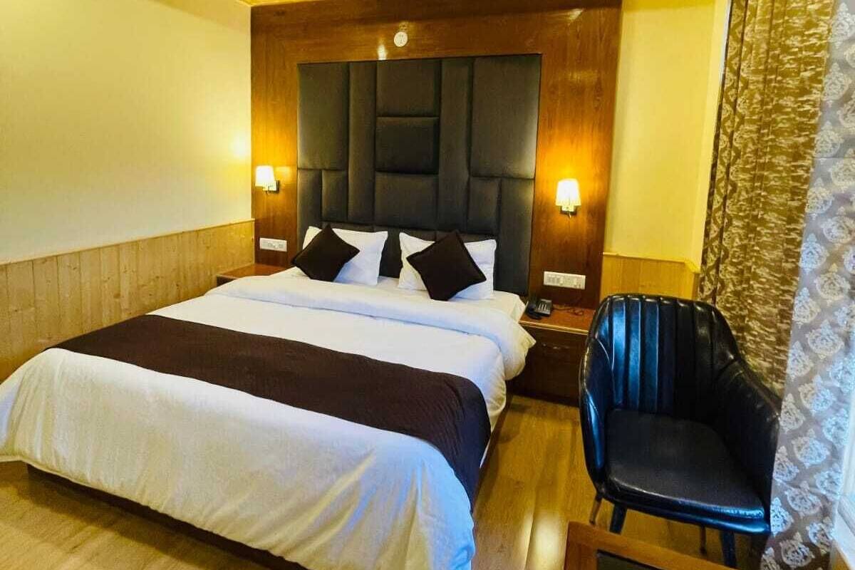 Quarto com teto de madeira, cama com roupa de cama branca e escura, uma cadeira preta e uma mesa de cabeceira com abajur.