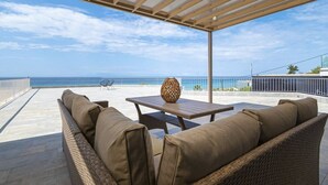A covered outdoor seating area with a view of the ocean, a wicker sofa, a table, and a decorative vase. - Althea Suites 4 by Rentals Pro - Nea Moudania Halkidiki (Nea Moudania)