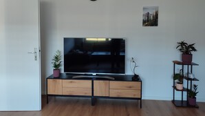 A modern living room with a flat-screen TV on a wooden cabinet, a door, a wall clock, a small framed picture, and a plant shelf. - Apartment 'Fewo Im Grünen Near Listertalsperre' with Private Terrace & Wi-Fi (Meinerzhagen)