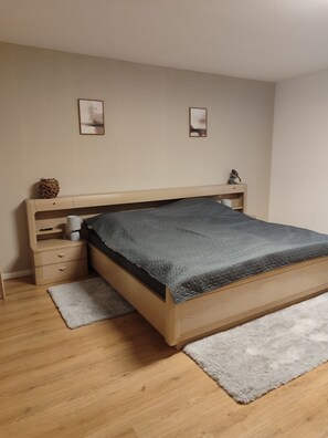 A bedroom with a wooden bed, a nightstand, a small shelf, and a gray rug. - Apartment 'Fewo Im Grünen Near Listertalsperre' with Private Terrace, Garden & Wi-Fi (Meinerzhagen)