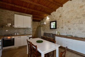 Une cuisine rustique avec des murs en pierre, des poutres apparentes en bois au plafond, une table à manger blanche et un four encastré.