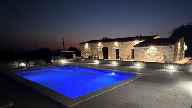 Une piscine éclairée de lumières bleues, un bâtiment en pierre doté de nombreuses fenêtres et une terrasse dallée.