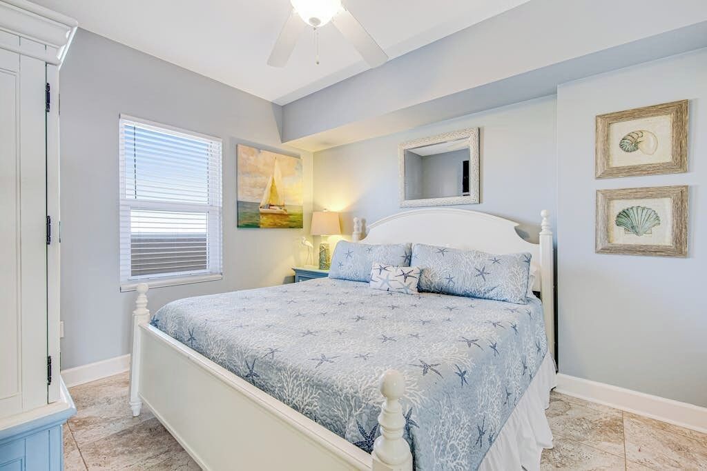 Ein Schlafzimmer mit Bett, Deckenventilator, Fenster mit Jalousien, Nachttisch mit Lampe und gerahmten Bildern an der Wand.