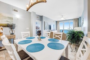 Un comedor con una mesa blanca, individuales azules, sillas blancas, un jarrón con flores y vista al océano.